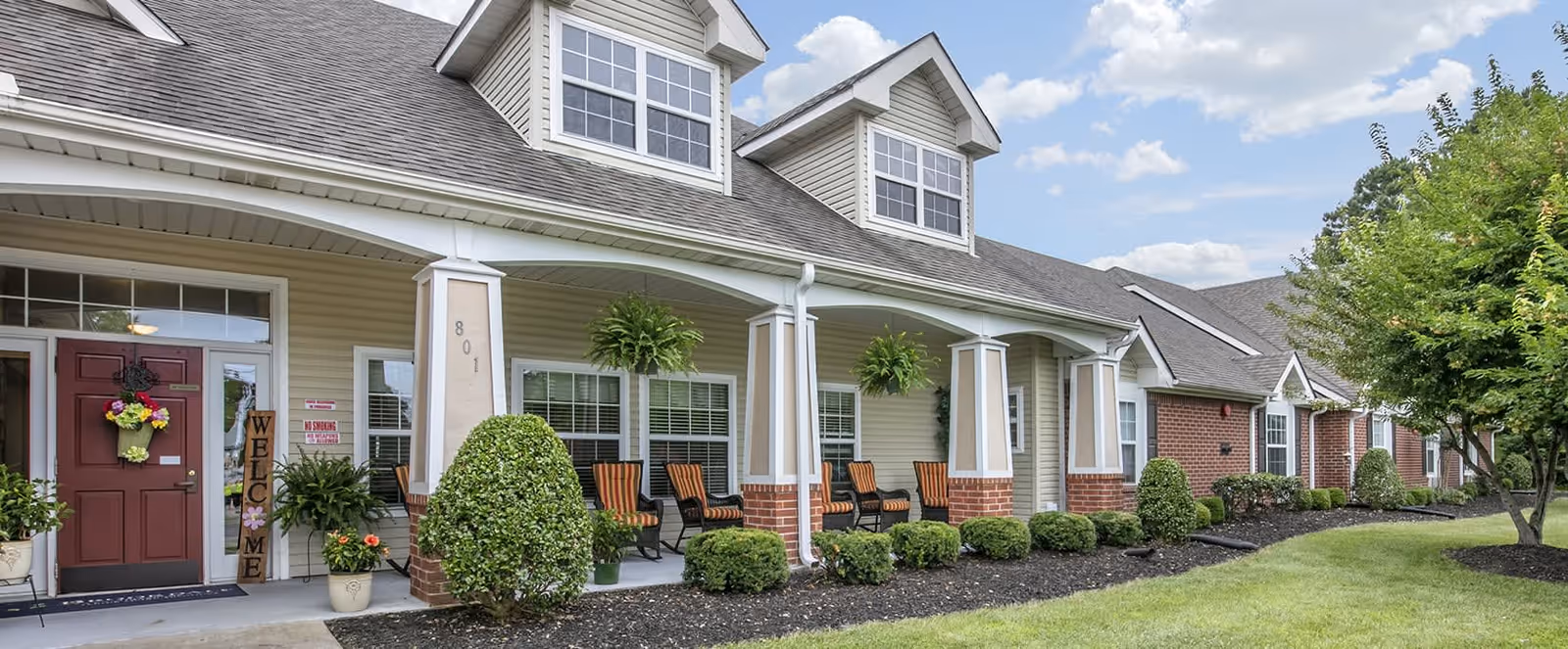 Exterior view of a senior living facility with a covered porch featuring several chairs, potted plants, and neatly trimmed bushes. The building has beige siding with brick accents and dormer windows on the roof under a partly cloudy sky.