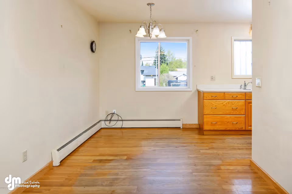 Empty room with wooden floor, beige walls, a window showing an outdoor view, a small kitchen counter with drawers and a sink on the right, a wall clock, and a ceiling light fixture with five bulbs.
