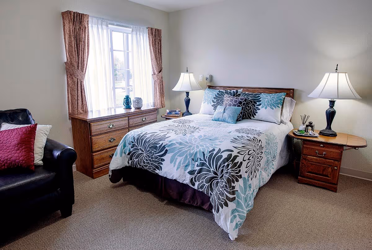 A cozy bedroom with a bed featuring a floral patterned comforter and multiple pillows. There are two bedside tables with lamps on each side of the bed. A wooden dresser is placed under a window with sheer white curtains and brown drapes. A black armchair with red and white pillows is positioned to the left of the dresser. The room has beige carpet flooring and light-colored walls.
