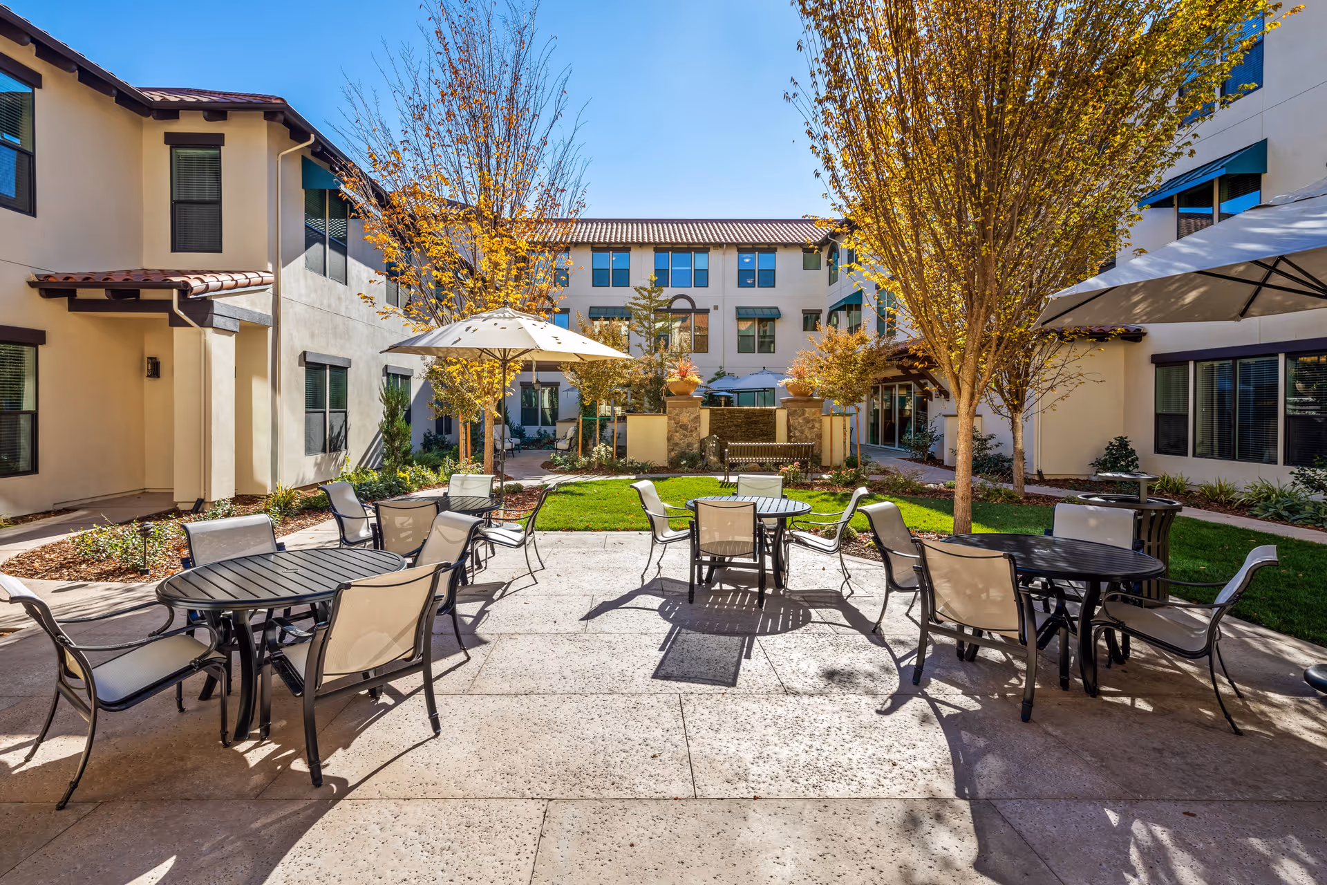 Outdoor courtyard area at Oakmont of East Sacramento with several round tables and chairs arranged on a paved patio. There are large umbrellas providing shade, trees with autumn foliage, and a multi-story building surrounding the courtyard under a clear blue sky.