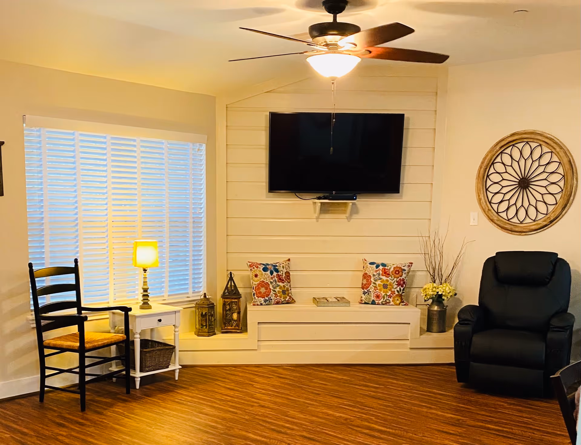 A cozy living room area with a ceiling fan and light fixture, a wall-mounted flat-screen TV above a built-in bench with two colorful floral pillows and decorative lanterns. To the left, there is a window with white blinds, a small white side table with a lit lamp, and a wooden chair with a woven seat. To the right, there is a black recliner chair and a round decorative wall piece above it. The floor is wood with a warm tone.