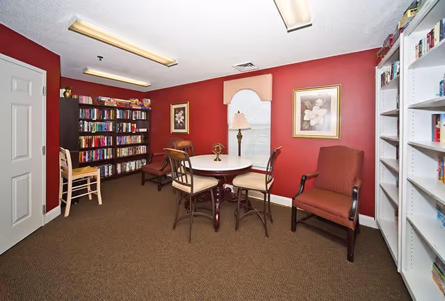 A cozy room with red walls featuring a round table with four chairs in the center. There are bookshelves filled with books along two walls, a floor lamp near a window with a valance, and framed floral artwork on the walls. The room has carpeted flooring and fluorescent ceiling lights.