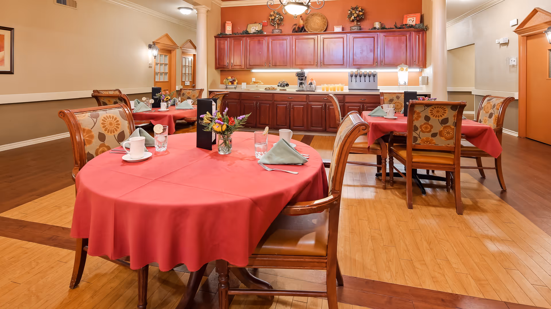 A dining room with round tables covered in red tablecloths, each set with cups, glasses, napkins, and small flower arrangements. The chairs have floral patterned upholstery. In the background, there is a wooden cabinet with a coffee machine and juice dispensers on the countertop. The room has warm lighting and wooden flooring.