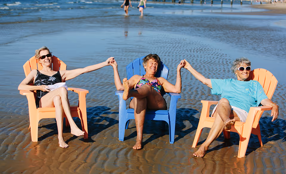 Three elderly women sitting on colorful Adirondack chairs on a sandy beach, holding hands and smiling, with shallow water and other beachgoers in the background.