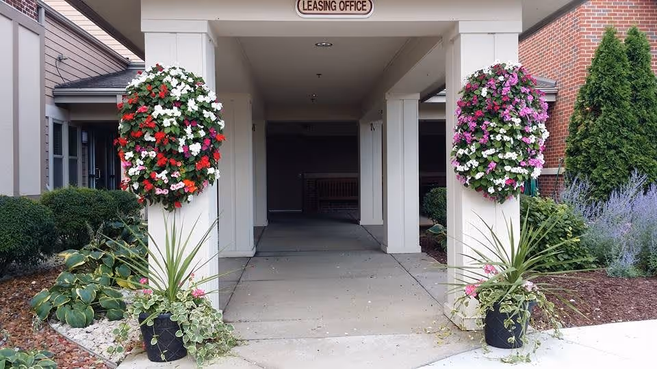 Entrance to a leasing office with a covered walkway supported by white pillars. Two large hanging flower baskets with red, white, and purple flowers are attached to the pillars. There are also potted plants with green foliage at the base of each pillar. Surrounding the entrance are landscaped bushes, plants, and a brick wall on the right side.