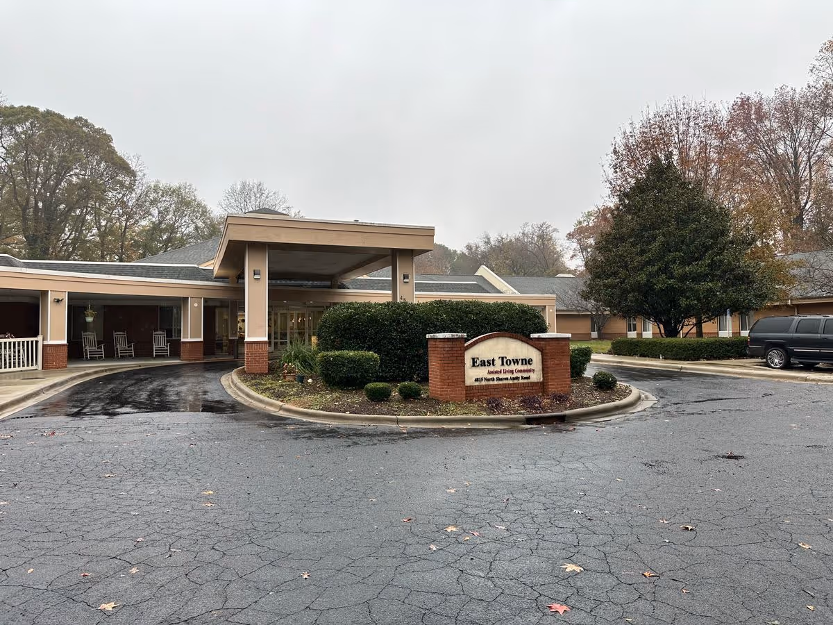 Front entrance and driveway of the East Towne assisted living community with a sign, covered porte-cochere, and surrounding landscaping on a cloudy day.