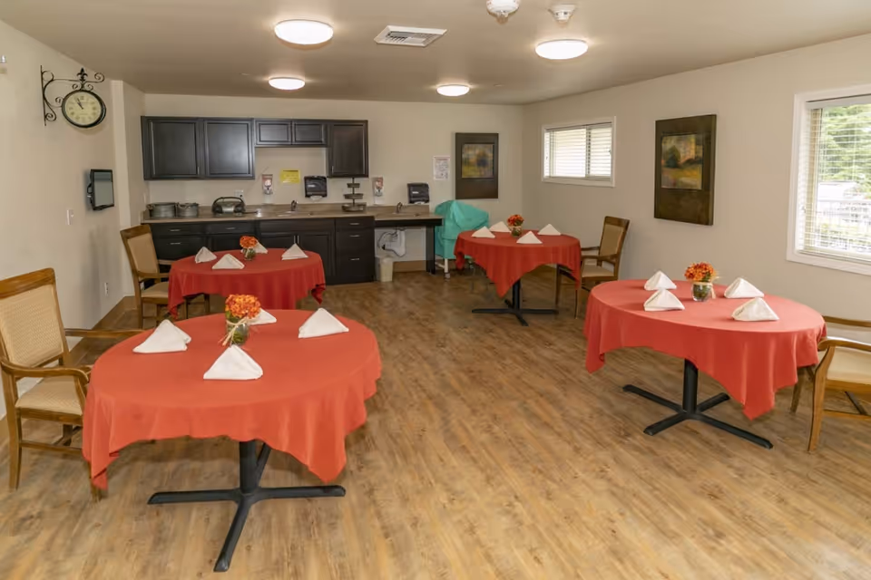 A dining room with four round tables covered with red tablecloths, each set with folded white napkins and small flower arrangements. The room has wooden flooring, beige walls, and several wooden chairs around the tables. There are two windows on the right wall letting in natural light, and a kitchenette with dark cabinets and a countertop along the back wall. A clock is mounted on the left wall.