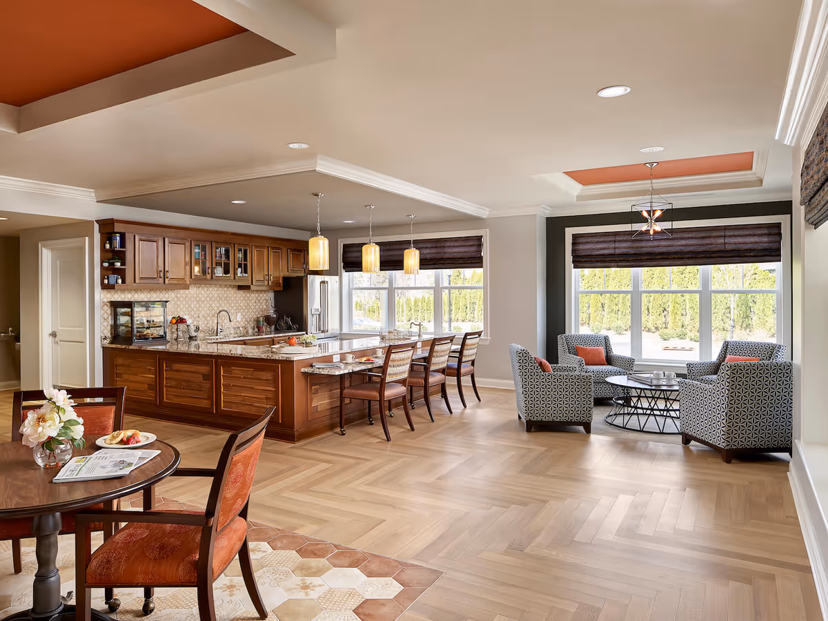 A spacious, well-lit interior of an assisted living facility featuring a kitchen area with wooden cabinets, a granite countertop island with bar stools, a small dining table with chairs, and a sitting area with four patterned armchairs around a round coffee table near large windows with brown blinds.