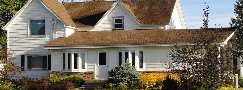 A two-story white house with brown shingled roof surrounded by trees and bushes under a clear blue sky.