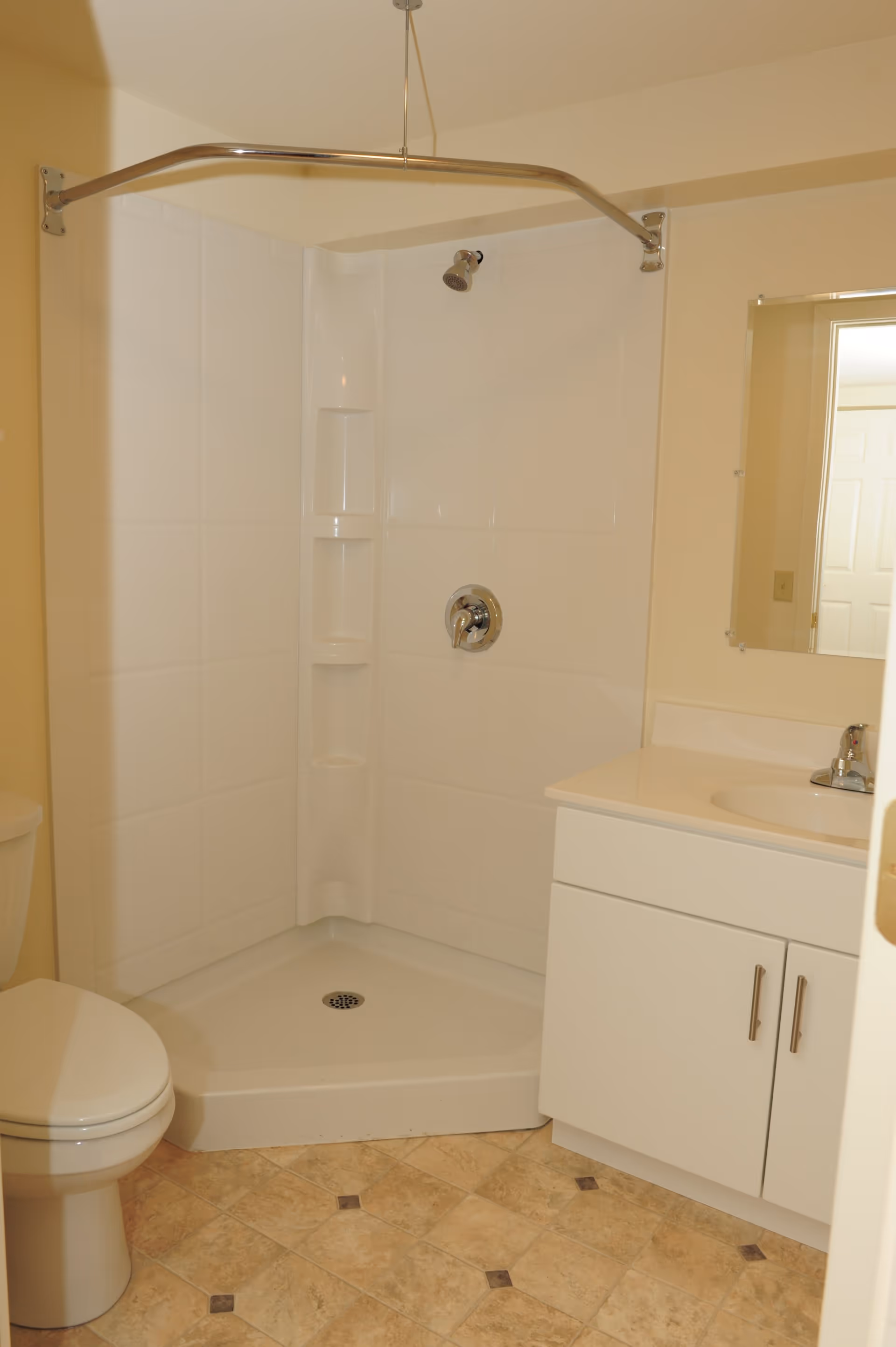 A small bathroom with a corner shower featuring white tiled walls and a chrome showerhead. There is a white toilet to the left and a white vanity with a sink and faucet to the right. A mirror is mounted on the wall above the vanity. The floor has beige and brown patterned tiles.