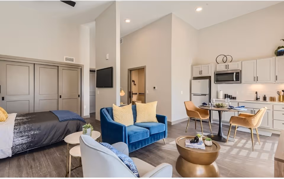 A modern, open-concept senior living apartment featuring a bedroom area with a bed and sliding closet doors, a living area with a blue loveseat and a gray armchair, and a kitchen with white cabinets, a refrigerator, microwave, and a round dining table with two tan chairs. The space is well-lit with natural light and has wood flooring.