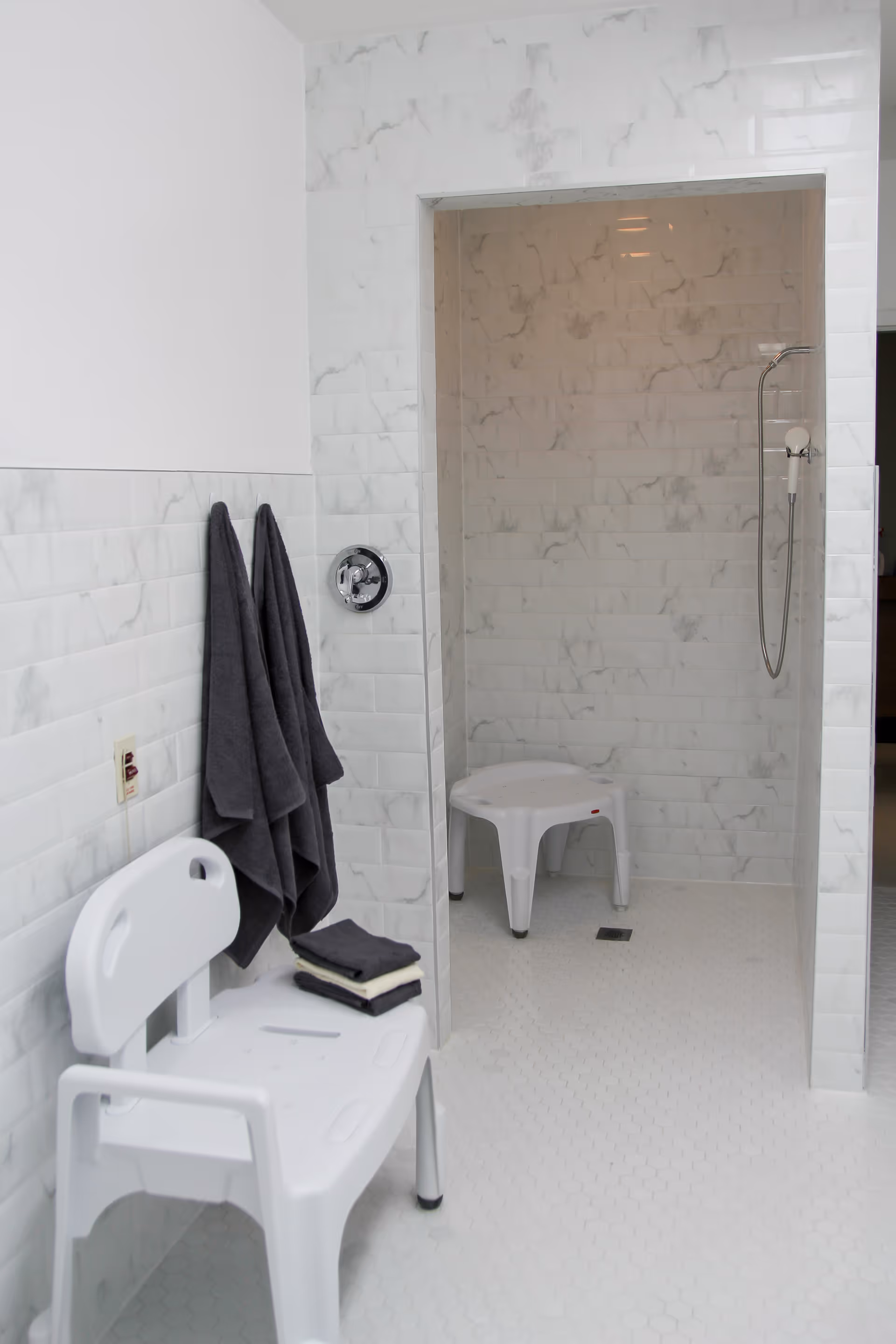 White tiled accessible shower area with two plastic shower seats, dark towels hanging on hooks, and a handheld shower head.