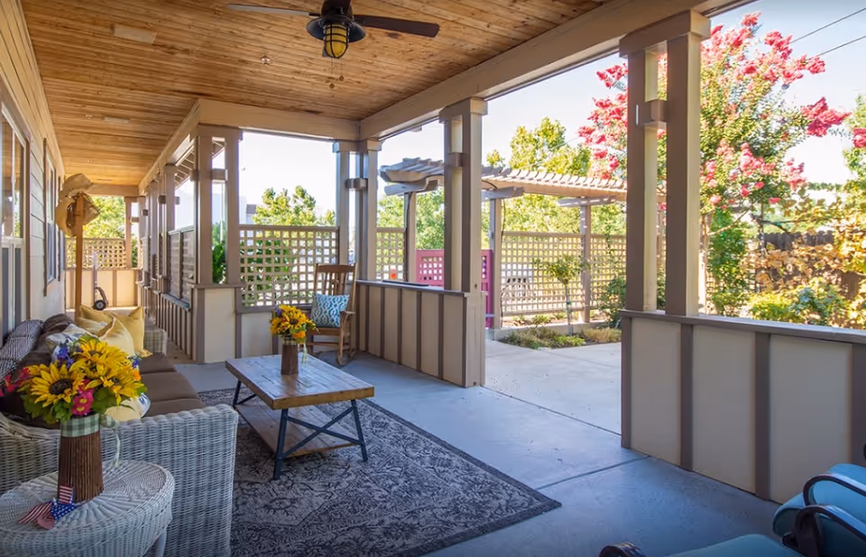 Covered outdoor patio area with wicker and cushioned seating, a wooden coffee table with a vase of sunflowers, ceiling fan, and lattice fencing. The patio overlooks a garden with trees and flowering plants under a clear blue sky.