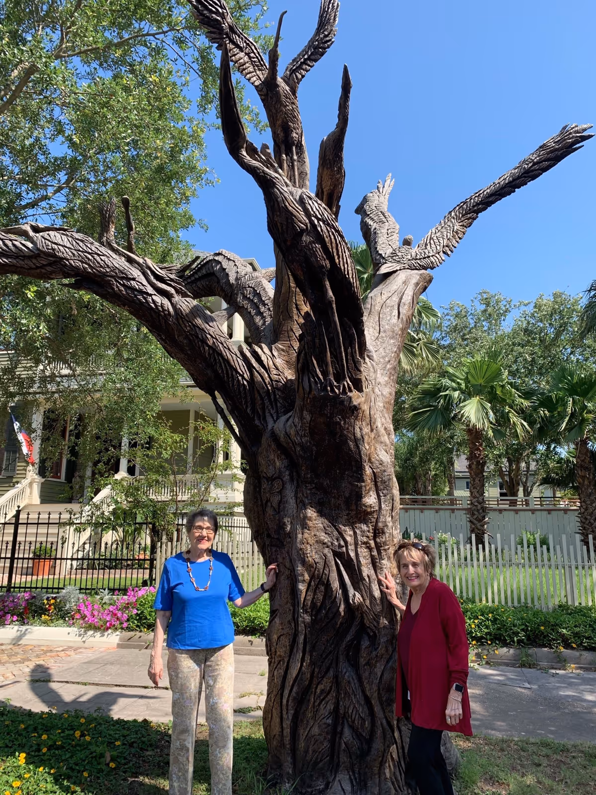 Two elderly women standing outdoors beside a large, intricately carved wooden tree sculpture with bird figures on its branches. Behind them is a white picket fence, palm trees, and a large house with a porch. The sky is clear and blue.