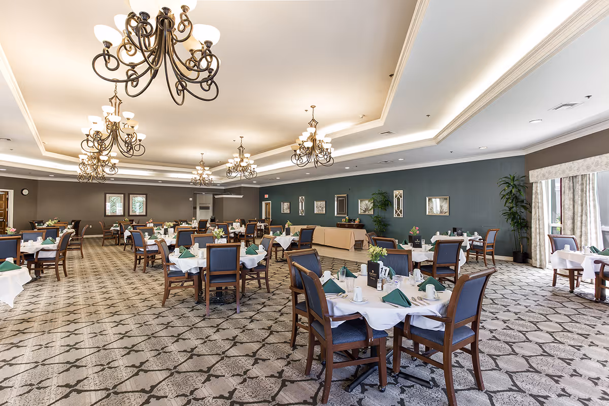 Spacious formal dining room with round tables set with white linens and green napkins beneath multiple ornate chandeliers.