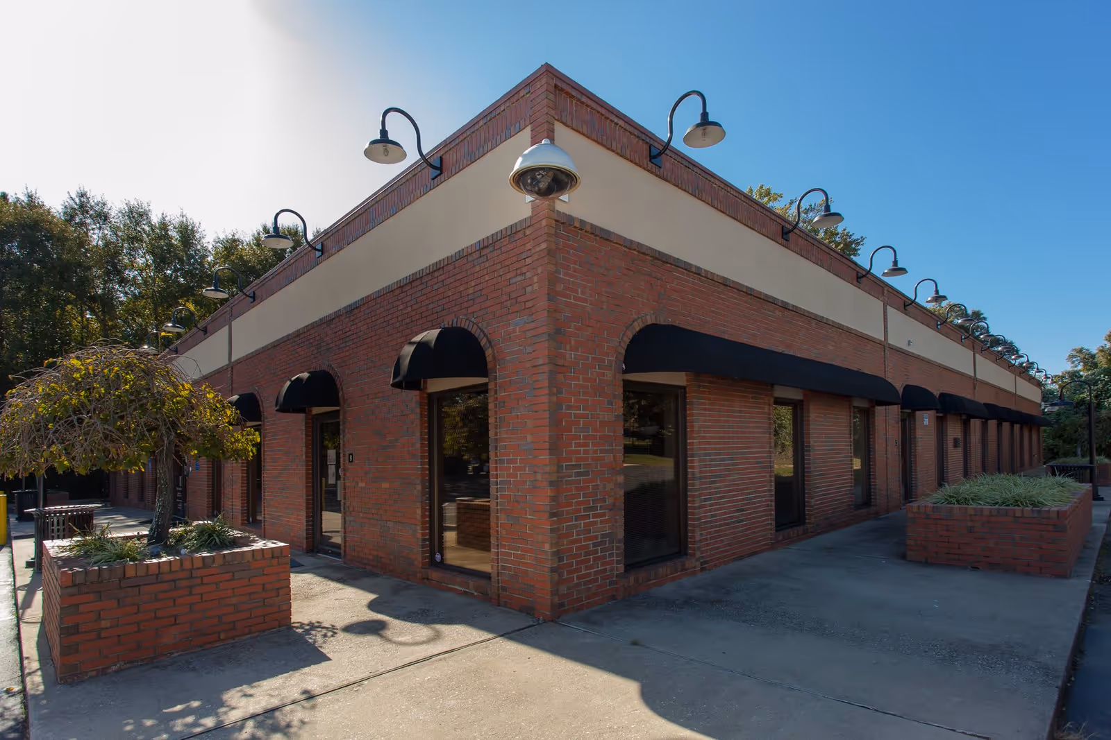 Exterior view of a single-story brick building with black awnings over windows and doors. The building has multiple outdoor wall-mounted lamps and a security camera at the corner. There are brick planters with greenery and a small tree near the building. The sky is clear and blue.