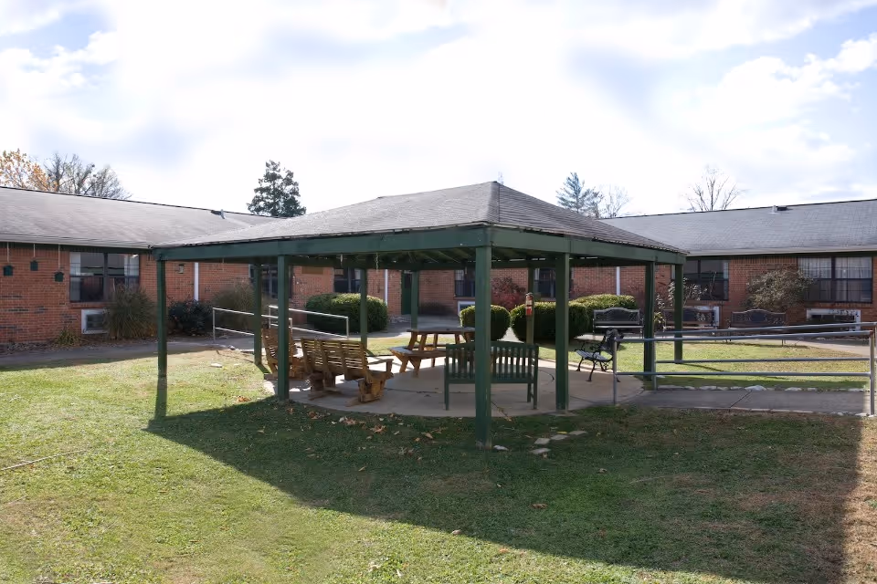 Outdoor courtyard area at Indian Creek Healthcare Center featuring a green metal gazebo with benches and picnic tables underneath. The courtyard is surrounded by a single-story brick building with windows and bushes. The sky is partly cloudy.