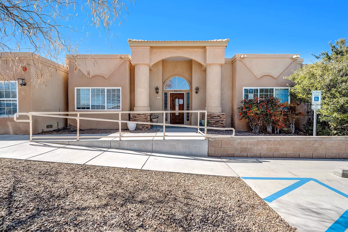 Front exterior view of Cottonwood Home, a single-story building with beige stucco walls, two large columns flanking the entrance, a wooden door with glass panels, and a wheelchair accessible ramp leading to the entrance. There are windows on either side of the door, some bushes and a reserved parking sign for handicapped parking visible on the right side.