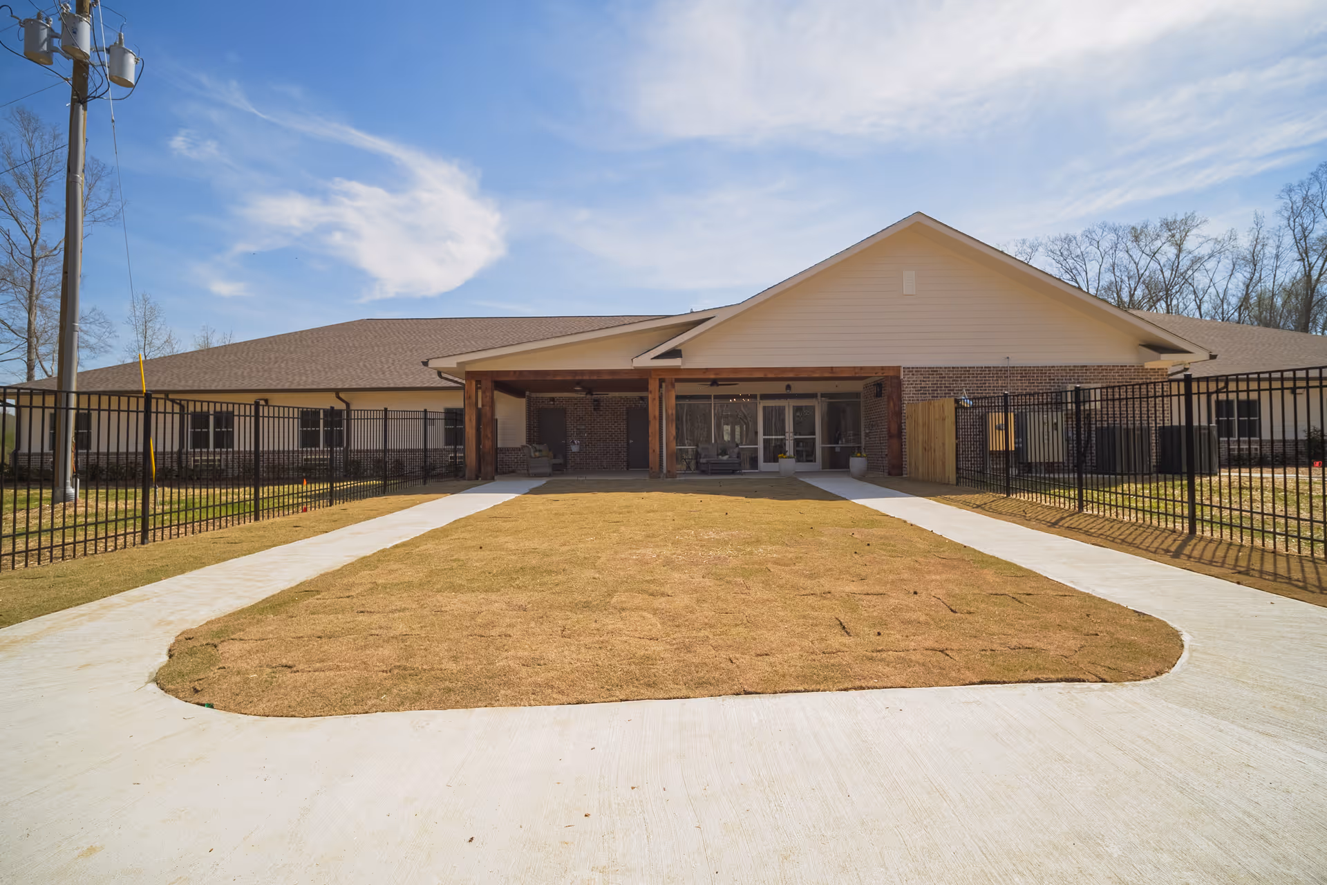 Front exterior view of a single-story building with a covered entrance supported by wooden beams, surrounded by a black metal fence and a concrete walkway leading to the entrance. The sky is clear with some clouds, and there are trees in the background.