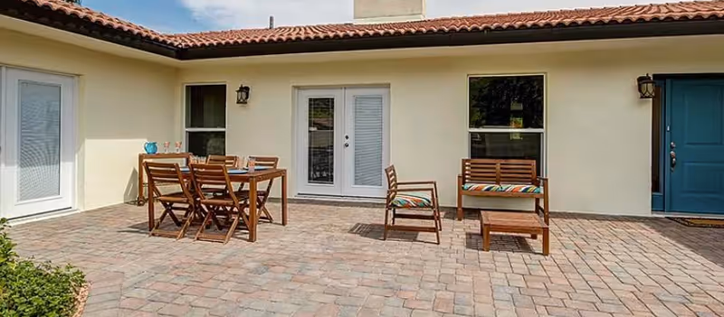 Outdoor patio area with a tiled floor, featuring a wooden dining table with six chairs on the left and a wooden bench with a cushion, a chair, and a small coffee table on the right. The patio is adjacent to a beige building with a blue door, two windows, and a set of white double doors.