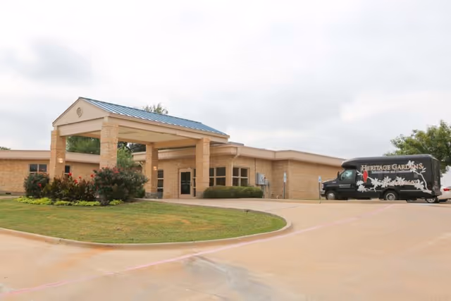 Exterior view of Heritage Gardens Rehabilitation & Healthcare building with a covered entrance supported by brick pillars, a landscaped area with bushes and flowers, and a black Heritage Gardens branded van parked on the driveway.