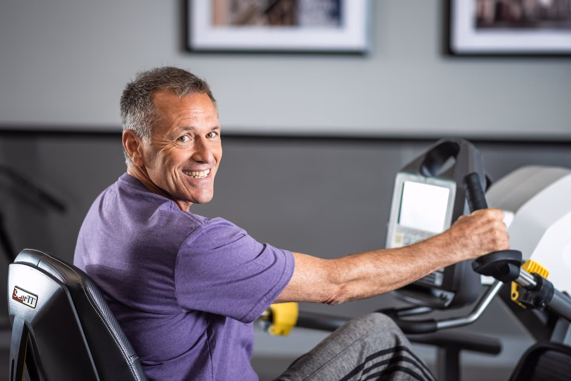A middle-aged man wearing a purple shirt is exercising on a seated fitness machine indoors, smiling and looking back towards the camera.