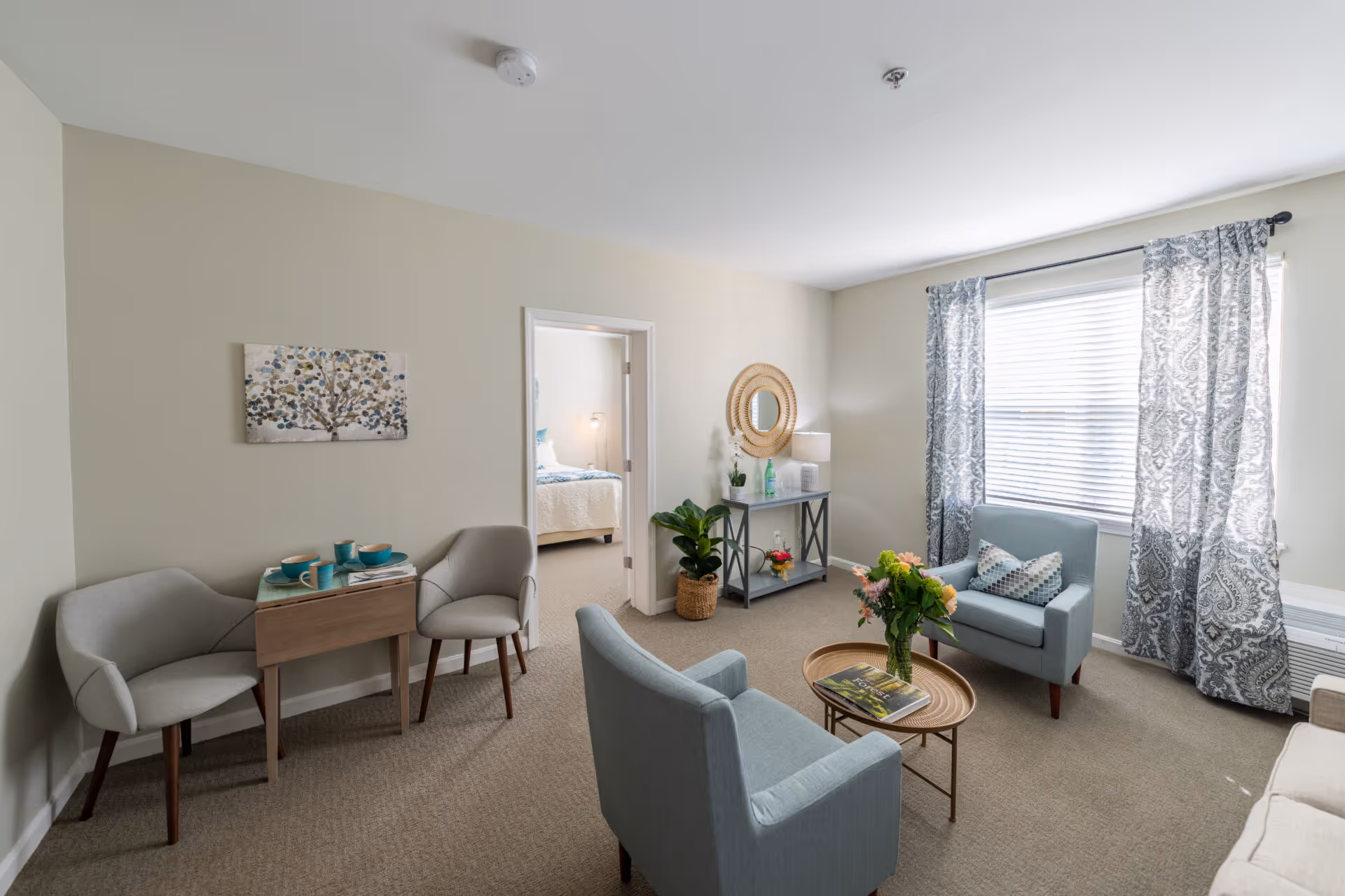 A cozy living room with two light blue armchairs around a small round wooden coffee table with a vase of flowers and a magazine. There is a window with patterned curtains letting in natural light. To the left, there are two gray chairs with a small wooden table set with teal cups and plates. A doorway leads to a bedroom with a bed visible. A console table with a round mirror, lamp, and decorative items is against the far wall.