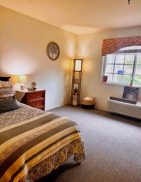 Sunlit furnished bedroom with a bed, nightstand, floor lamp, window and a wall air-conditioning unit.