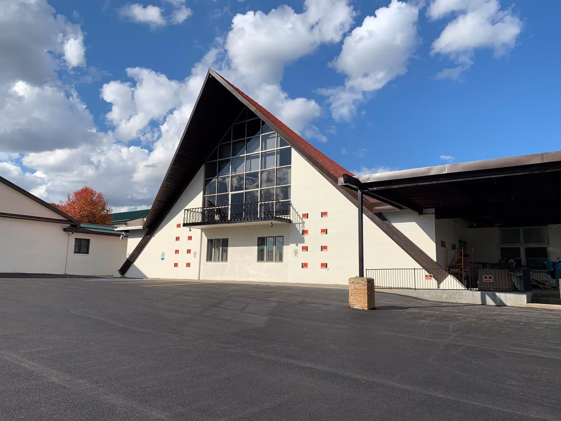 Exterior view of a building with a steep triangular roof and large glass windows reflecting the sky and clouds. The building has white walls with red accents and a paved parking area in front under a partly cloudy blue sky.