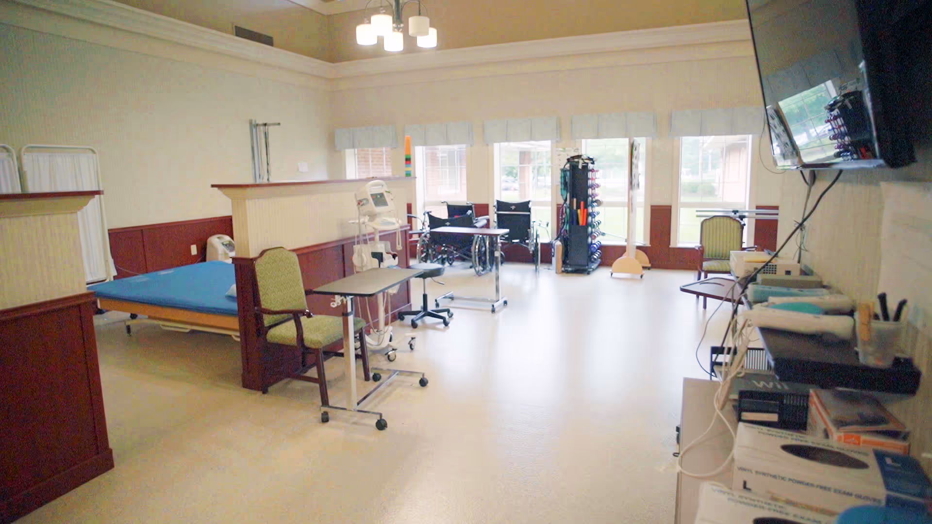Rehabilitation room with a bed, chairs, wheelchairs, therapy equipment and large windows.