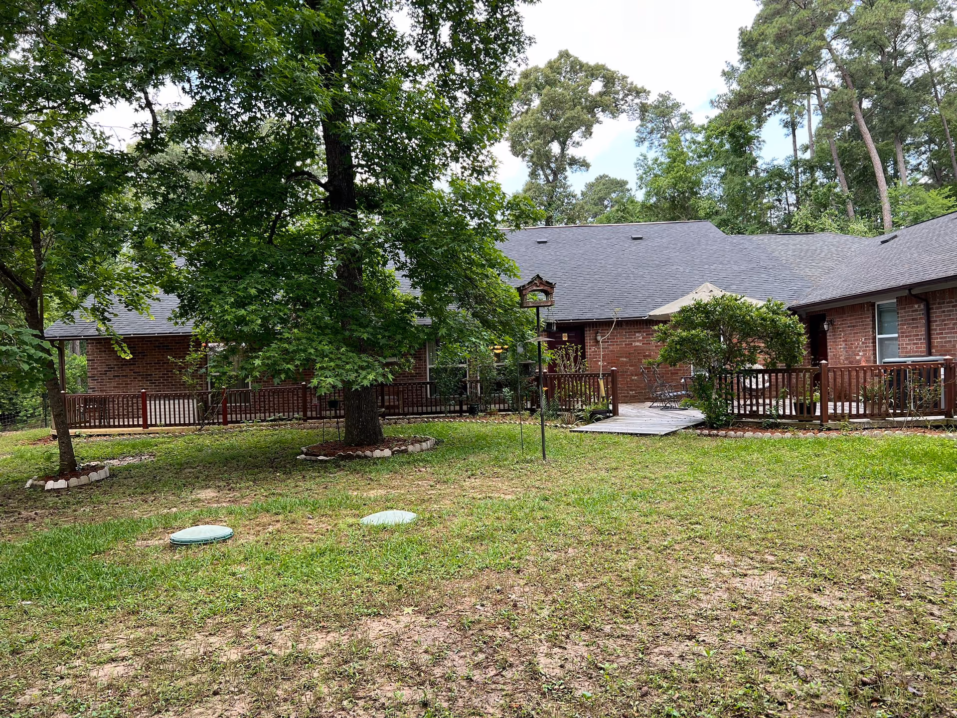 Outdoor view of a senior care facility with a brick building surrounded by trees and greenery. There is a wooden deck with patio furniture and an umbrella, and a bird feeder is visible near the center. The area has a grassy lawn with some patches of dirt.