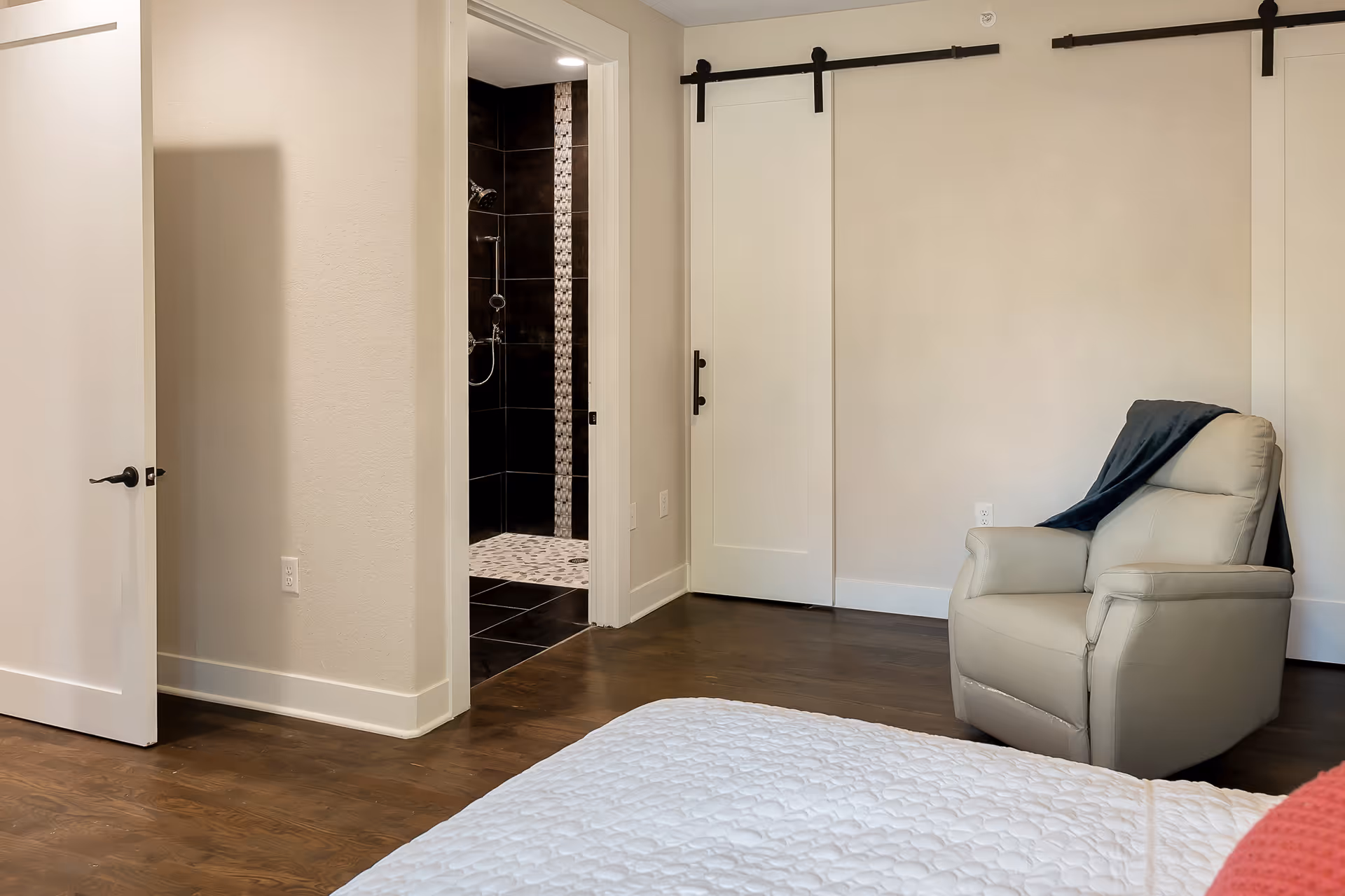 A bedroom with a white quilted bedspread partially visible in the foreground, a beige recliner chair with a dark throw draped over it, light-colored walls, dark hardwood floors, a white sliding barn door, and an open door leading to a bathroom with dark tiles and a mosaic tile shower floor.