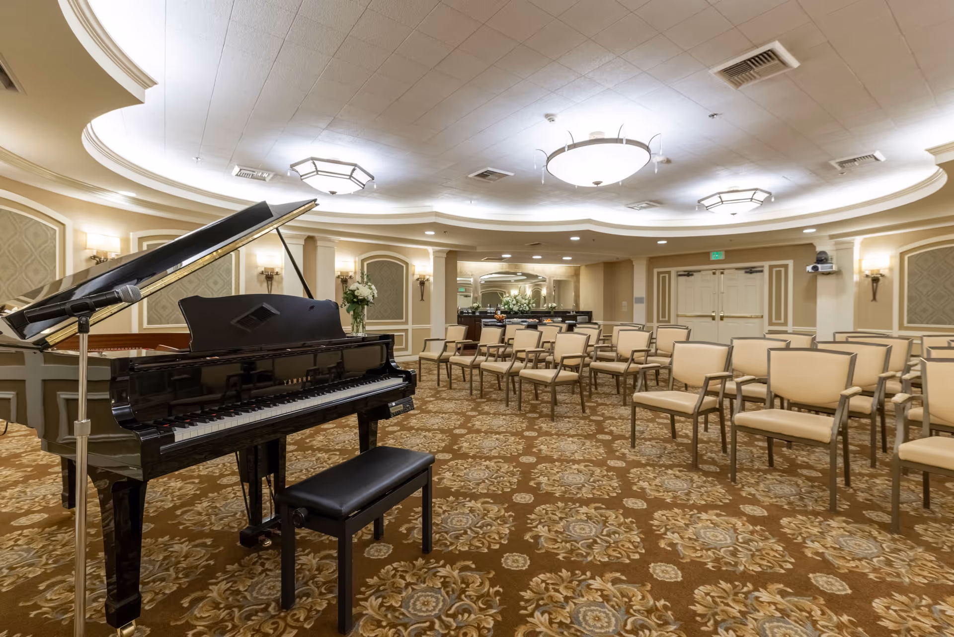 A spacious room with a grand piano and a microphone stand on the left side, rows of beige cushioned chairs arranged facing the piano, ornate patterned carpet, elegant wall sconces, and a large circular ceiling light fixture in the center.