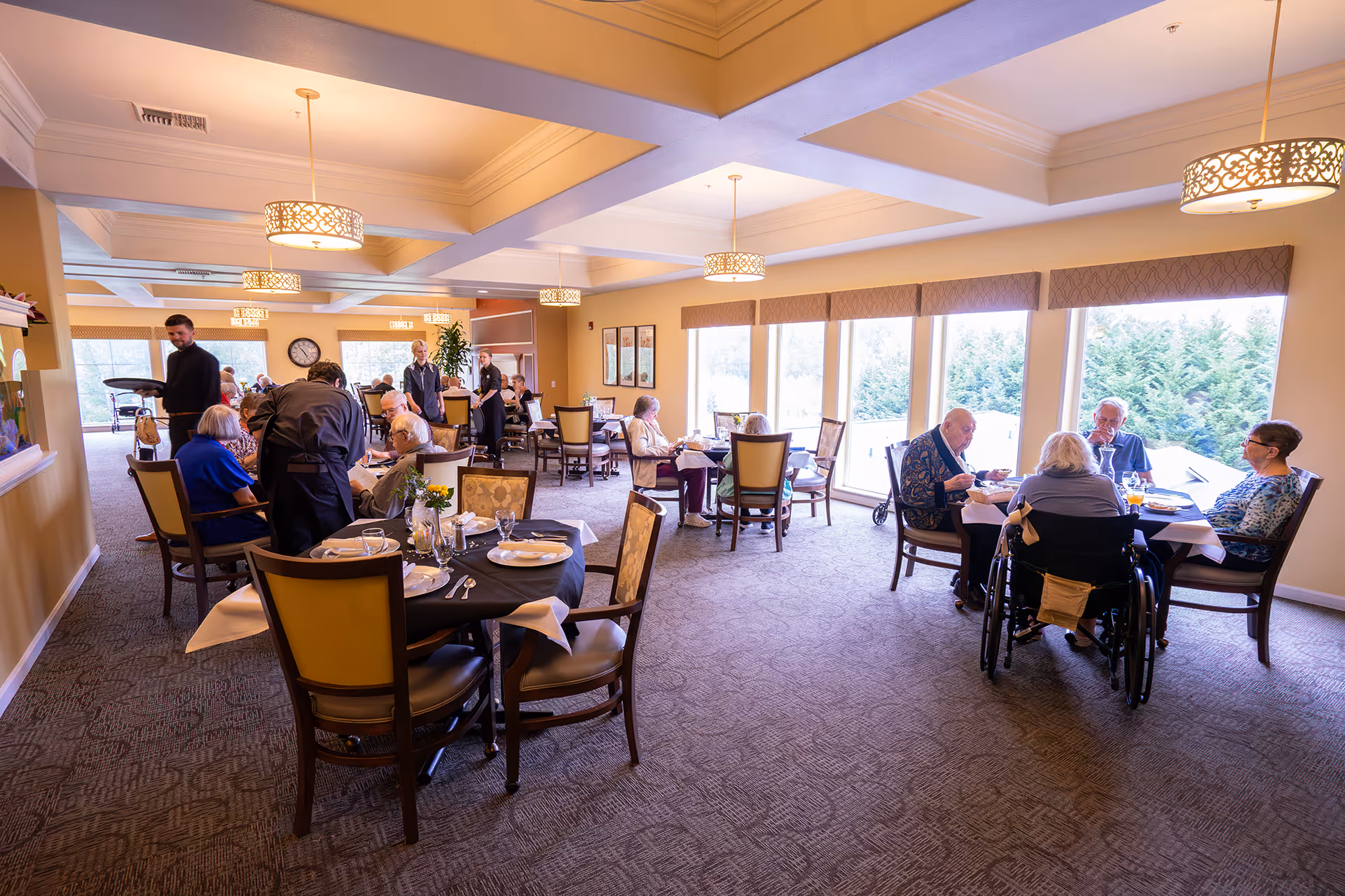 A bright dining room in a senior living facility with several elderly residents seated at tables enjoying their meals. The room has large windows letting in natural light, decorative hanging light fixtures, and staff members attending to the residents.