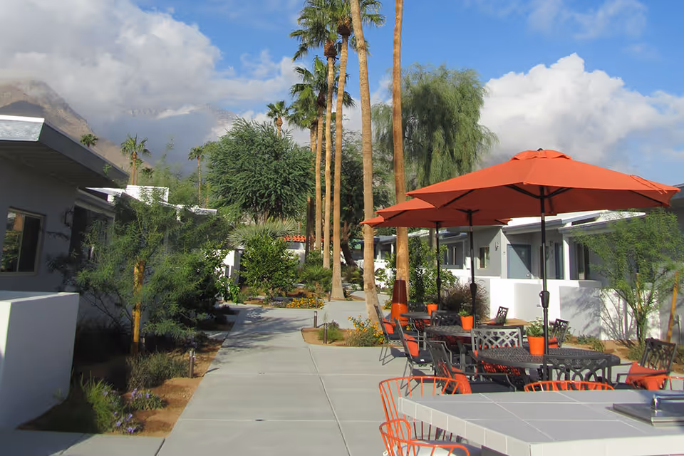 Sunny courtyard with palm trees, a paved walkway and outdoor seating under orange umbrellas between low-rise buildings.