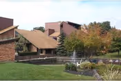 Exterior view of Williamstowne Senior Apartments showing a building with a sloped roof, surrounded by green grass, trees with autumn foliage, a pond with a fountain, and a small bridge over the water.