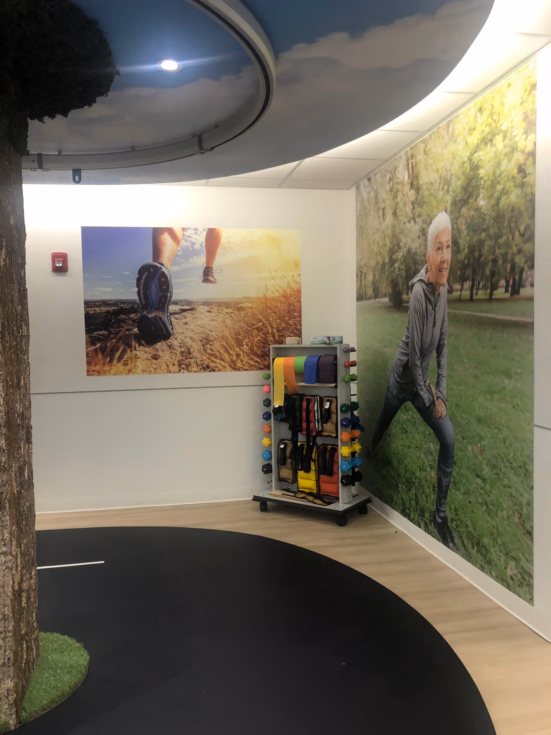 Indoor exercise area with a black circular mat on the floor, a tree trunk feature, and a rack holding colorful exercise bands and weights. The walls have large photos of a person running on a trail and an elderly woman stretching outdoors in a park.