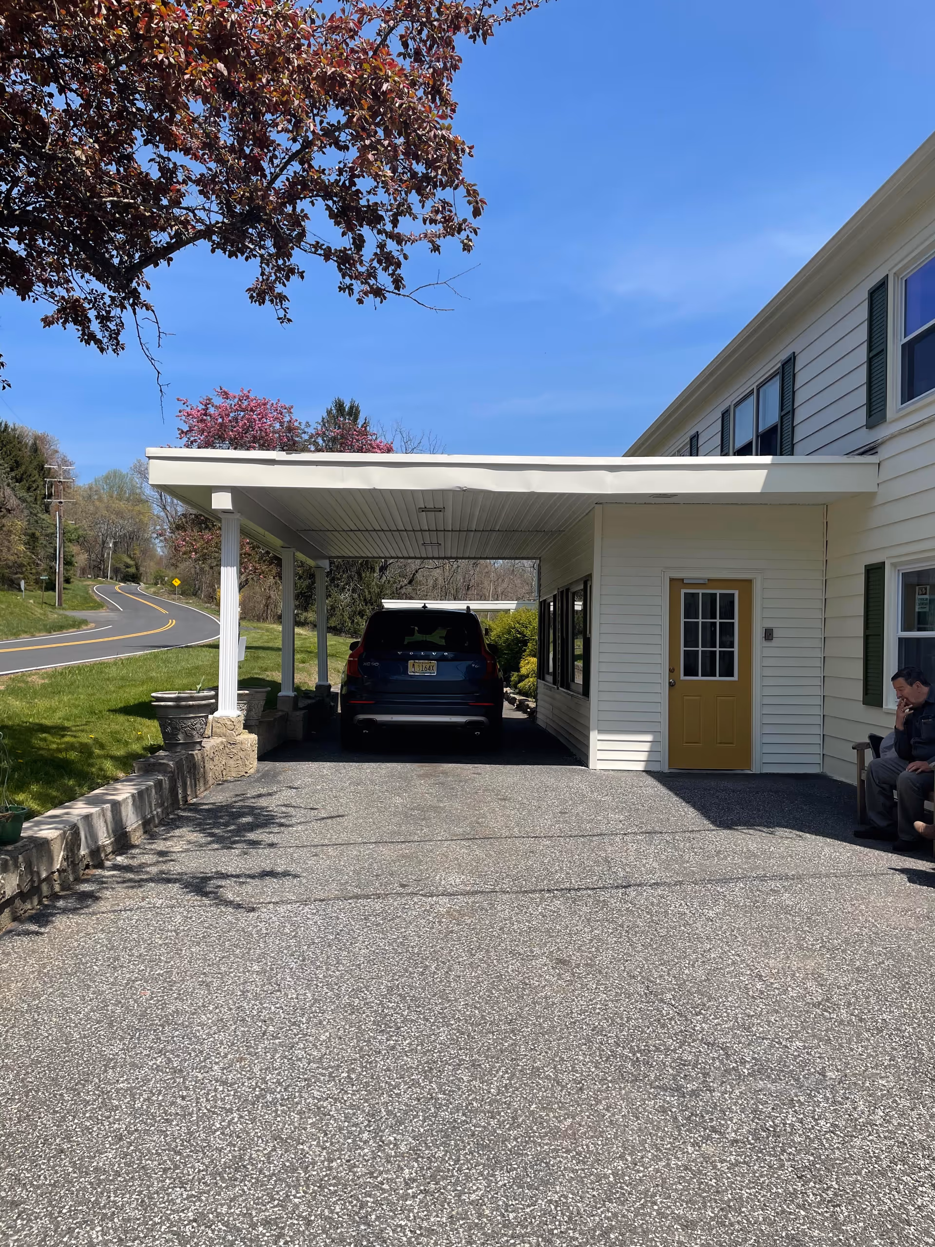 A driveway with a covered carport attached to a two-story white building with green shutters and a yellow door. A black SUV is parked under the carport. There is a stone retaining wall on the left side of the driveway and a man sitting on a bench near the building on the right. Trees with spring foliage and a clear blue sky are visible in the background.