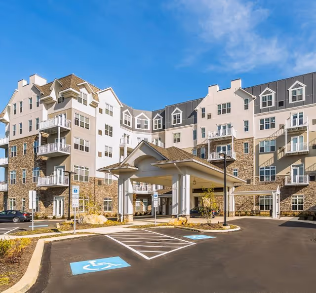 Exterior view of a multi-story senior living facility with stone and light-colored siding, featuring balconies, a covered entrance, and accessible parking spaces in front under a clear blue sky.