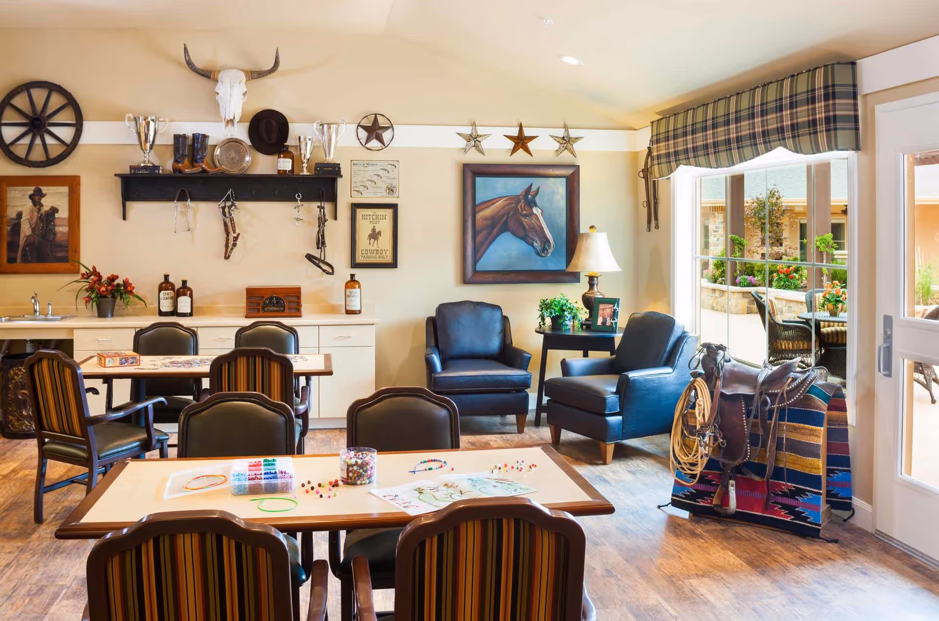 A cozy common area in a memory care community decorated with a western theme, featuring a table with chairs and craft supplies, two leather armchairs with a side table and lamp, western decor on the walls including a horse painting, cowboy hat, boots, and a skull. Large windows with plaid valances let in natural light and show an outdoor patio with seating.