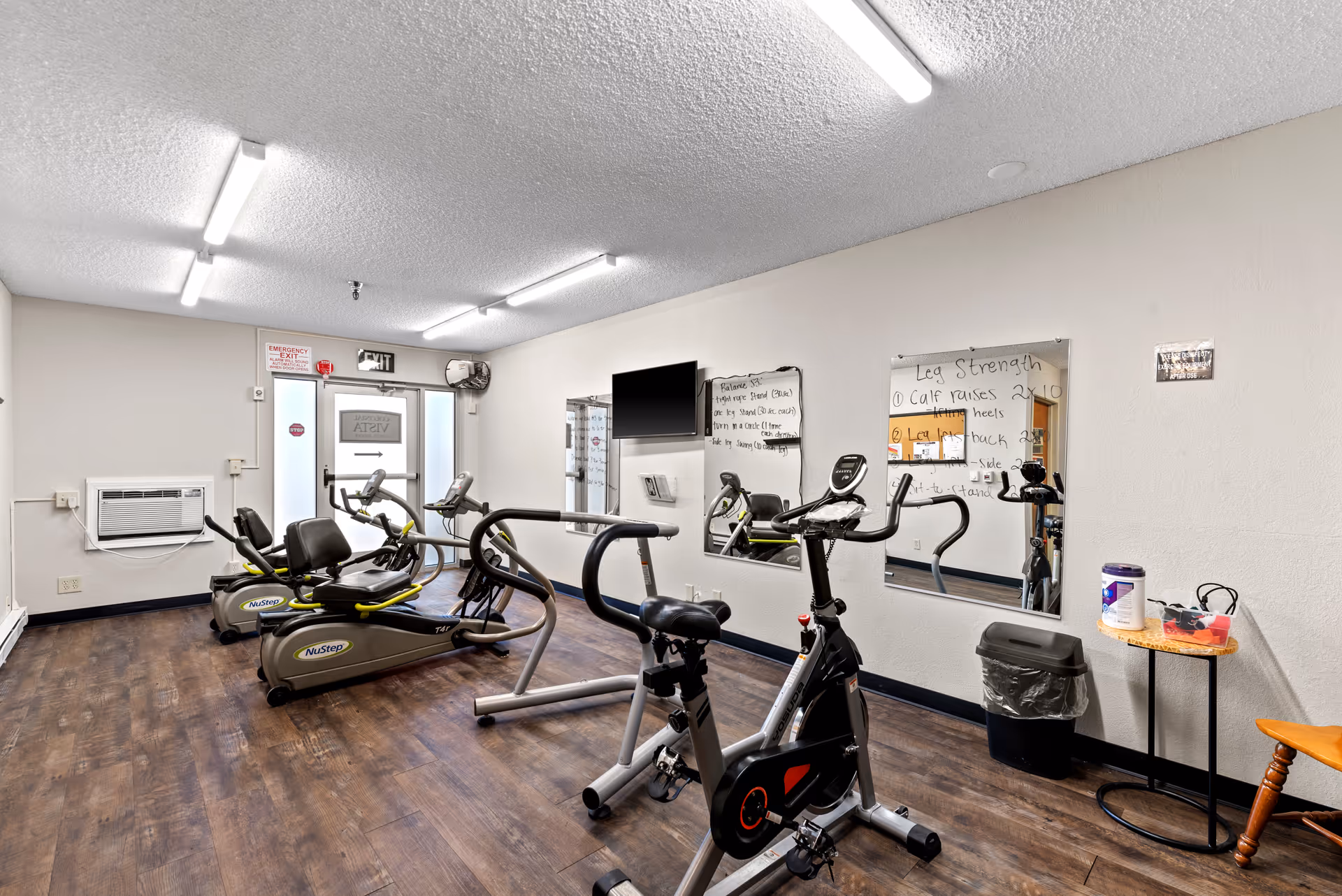 Small fitness room with stationary bikes, recumbent steppers, mirrors, whiteboards and wood-look flooring.