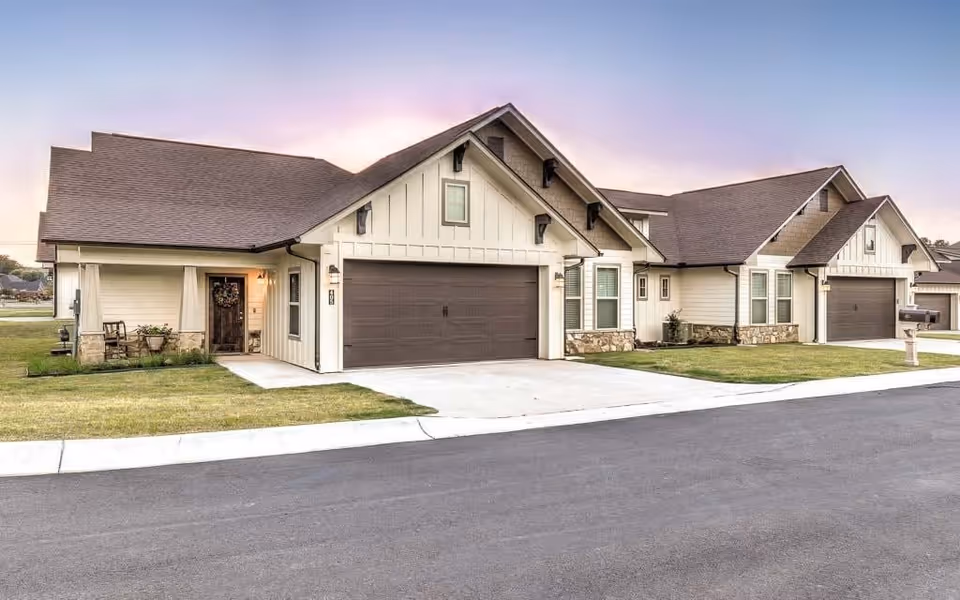 Front view of single-story attached homes with garages, driveways, and lawns under a twilight sky.