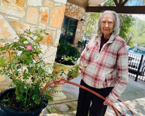 An elderly woman wearing a plaid jacket is watering a potted plant with pink flowers outside a stone building under a covered patio.