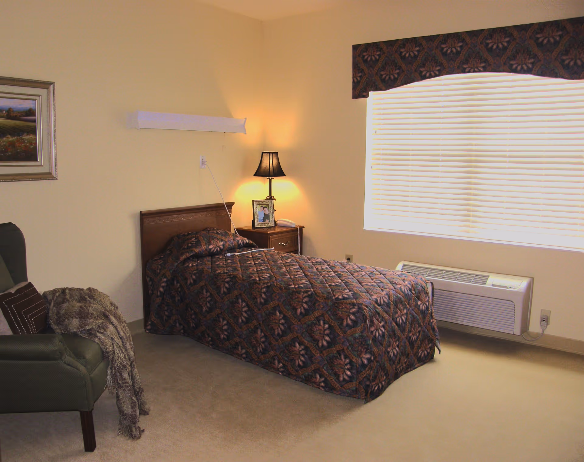 A bedroom with a single bed covered in a dark patterned quilt, a wooden nightstand with a lamp and a framed photo, a green armchair with a blanket draped over it, a large window with closed blinds and a decorative valance, and a wall-mounted air conditioning unit below the window.