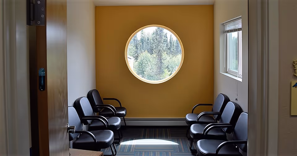 A small waiting room with two rows of black chairs facing each other. The back wall is painted yellow and features a round window showing trees outside. There is a rectangular window with blinds on the right wall, and the door to the room is partially open.