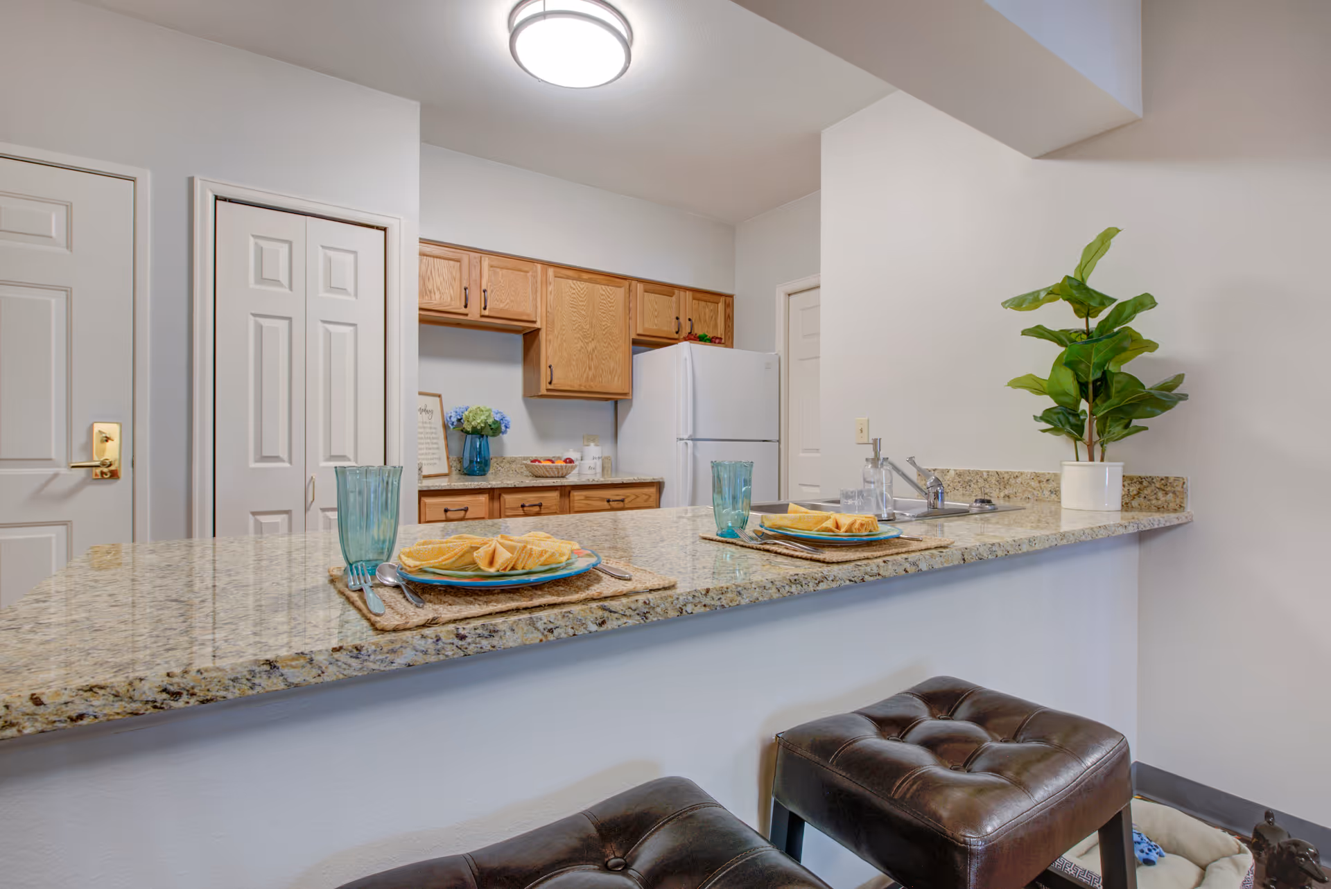 Granite breakfast bar with place settings and stools in front of a kitchen with wooden cabinets and a refrigerator.