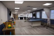 Interior view of a spacious therapy or activity room with exercise equipment, therapy tables, a walker, and a rack of colorful balls and toys along the left wall. The room has large windows with curtains, light wood flooring, and ceiling lights.
