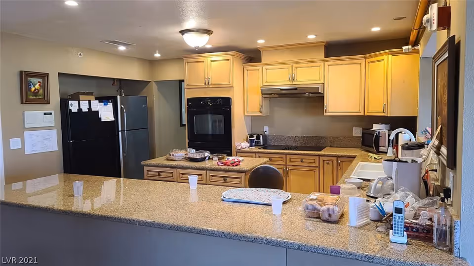 Interior view of a kitchen with beige wooden cabinets, a black oven, a stainless steel refrigerator, and a granite countertop island. Various kitchen items including a cordless phone, coffee maker, and food containers are placed on the countertops.