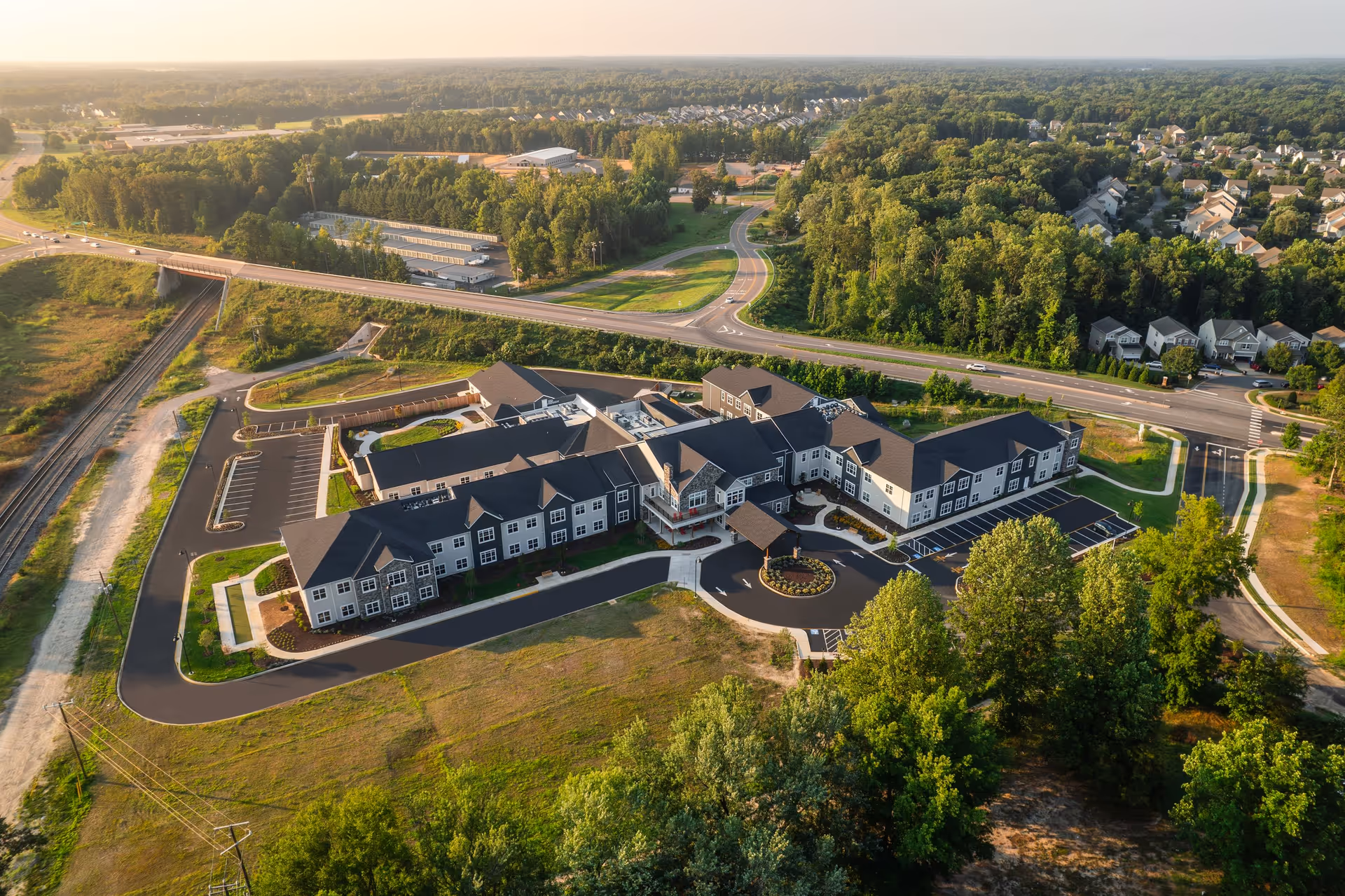 Aerial view of Sancerre Atlee Station, a large senior living facility surrounded by greenery, roads, and nearby residential neighborhoods. The building has a modern design with multiple wings, a circular driveway at the entrance, and ample parking spaces. The surrounding area includes trees, open fields, and a highway overpass.