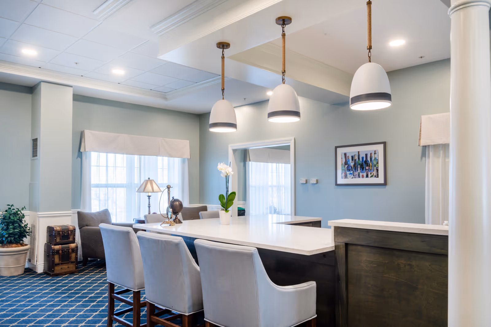 Bright and spacious common area with a large white countertop island surrounded by cushioned chairs. Three modern pendant lights hang above the island. The room features light blue walls, a patterned blue carpet, a potted plant, a small table with a lamp and decorative items, and a framed artwork on the wall. Large windows with white curtains allow natural light to fill the space.
